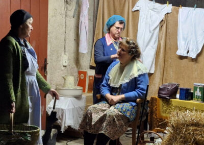 Historische Theaterszene mit drei Personen in traditioneller Kleidung, eine Person sitzt auf Stuhl, Wäsche hängt im Hintergrund, rustikale Atmosphäre mit Stroh am Boden