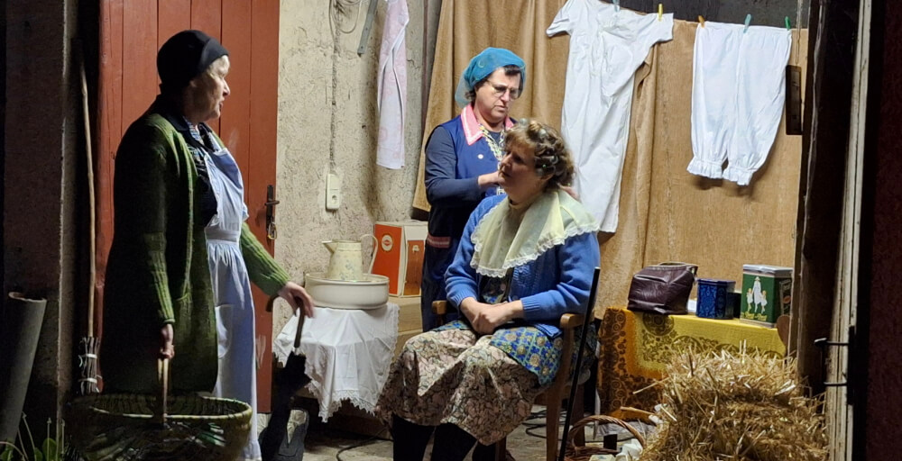 Historische Theaterszene mit drei Personen in traditioneller Kleidung, eine Person sitzt auf Stuhl, Wäsche hängt im Hintergrund, rustikale Atmosphäre mit Stroh am Boden