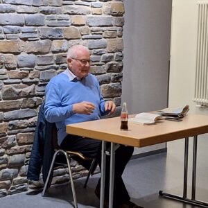 Werner Lutz am Tisch bei der Lesung, Älterer Mann in hellblauem Pullover hält Lesung an Tisch mit aufgeschlagenem Buch vor Natursteinwand